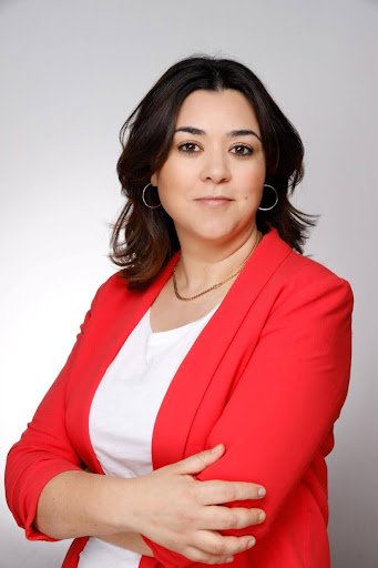 Frau mit dunklem, schulterlangem Haar, weißem Oberteil und rotem Blazer schaut frontal in die Kamera